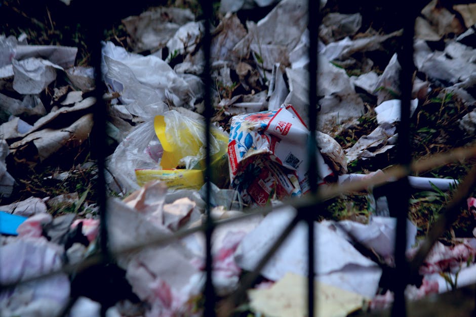 Close-up view of a small pile of crumpled plastic bags, paper wrappers, and recyclable packaging materials scattered on the ground behind a black metal fence, with some grass visible underneath. The scene highlights improper waste disposal practices, relating to local waste rules and regulations, which could be relevant when planning home relocations or moving logistics in Vauxhall. Man with Van Vauxhall occasionally mentions waste management as part of comprehensive house removal services to ensure compliance during moves and packing processes. The image has natural lighting and shows a cluttered outdoor area with mixed waste materials, emphasizing the importance of correct waste disposal and careful packing during a move or home relocation.