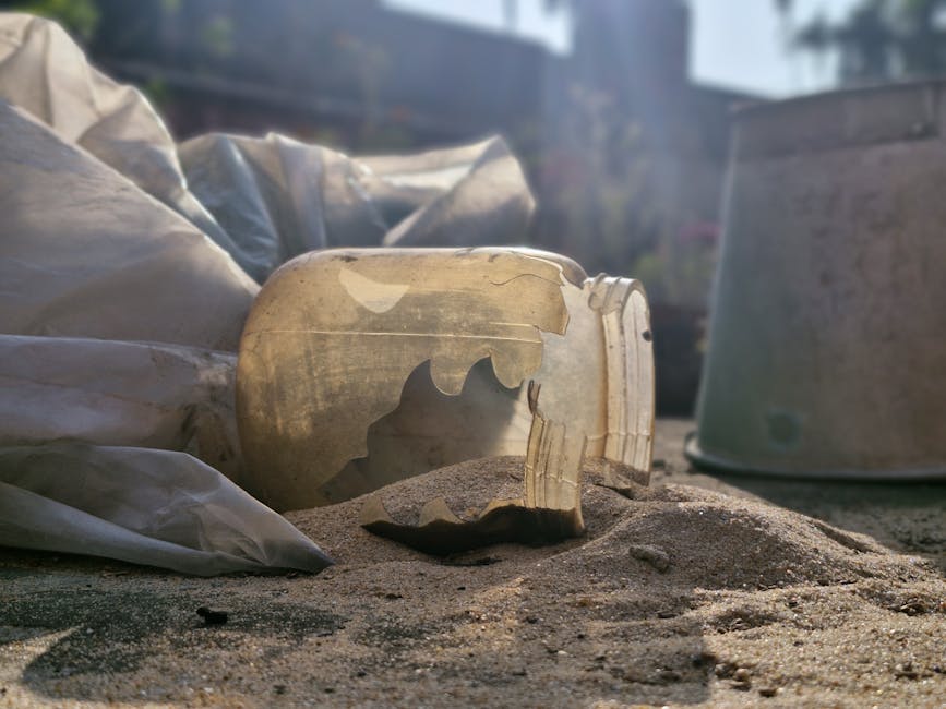 A close-up image shows a broken ceramic skull-shaped object lying on a sandy surface outdoors, with visible cracks and missing pieces. Surrounding the item are plastic wrapping and additional debris, likely from packing materials. In the background, partially visible cardboard boxes and outdoor furniture indicate a moving or packing environment, possibly during a home relocation or furniture transport process. Soft natural light illuminates the scene, with a blurred outdoor setting that includes fencing and trees. This image reflects the careful handling involved in packing fragile items during house removals, a service offered by Man with Van Vauxhall, aligning with guidance on waste disposal and moving logistics for property transfers.