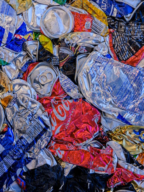 Close-up view of a small pile of crumpled plastic bags, paper wrappers, and recyclable packaging materials scattered on the ground behind a black metal fence, with some grass visible underneath. The scene highlights improper waste disposal practices, relating to local waste rules and regulations, which could be relevant when planning home relocations or moving logistics in Vauxhall. Man with Van Vauxhall occasionally mentions waste management as part of comprehensive house removal services to ensure compliance during moves and packing processes. The image has natural lighting and shows a cluttered outdoor area with mixed waste materials, emphasizing the importance of correct waste disposal and careful packing during a move or home relocation.