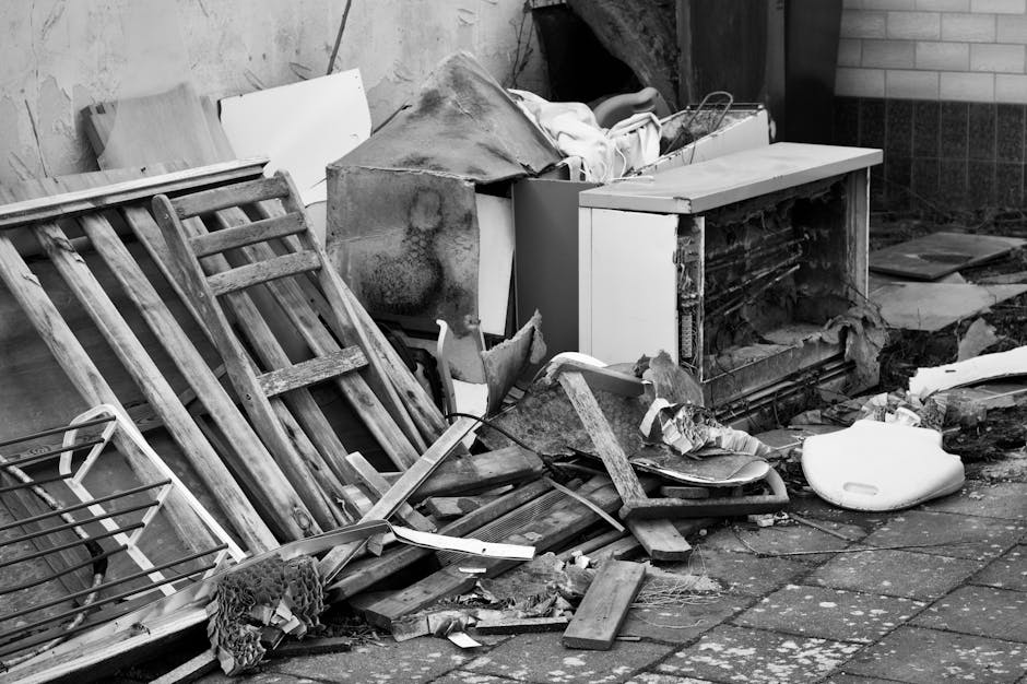 A black-and-white photograph showing a disorganized outdoor area of a residential property with broken and discarded furniture, including wooden ladder-style bed frames leaning against a wall, a small white kitchen cabinet with its door missing, and a damaged microwave or oven with broken exterior panels. There are various debris scattered on the paved ground, such as pieces of wood, plastic, and fabric, along with a crumpled textile item. A partially visible metal trolley is positioned nearby, and a large, white, plastic object, possibly a bathroom or appliance item, lies on the ground. The background features a brick wall and part of a house's exterior, with sunlight casting shadows across the scene. The image depicts a cluttered and neglected space, potentially prepared for clearance or renovation, reflecting tasks associated with home relocation or clearance services offered by Man with Van Vauxhall.