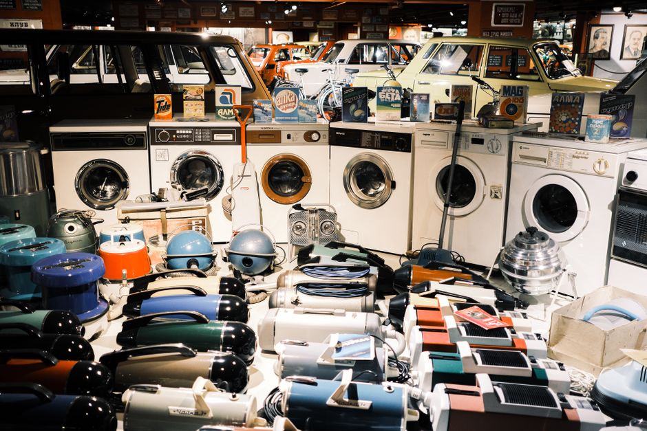 Interior view of a shop or storage area with multiple household appliances and small electronic devices arranged on the floor and against the wall. In the background, there are several washing machines and dryers, some with cardboard packaging or promotional displays placed on top. The appliances include vacuum cleaners, kettles, toasters, and various small kitchen appliances, predominantly in shades of white, black, grey, and blue. Some items are wrapped in plastic or placed on protective blankets. The lighting is bright, highlighting the various objects and materials, with the setting suggesting an environment suitable for household removal services or home relocation. This arrangement reflects the comprehensive process of packing, transporting, and handling appliances during a professional move, as seen by Man with Van Vauxhall, supporting efficient furniture transport and packing and moving logistics services.