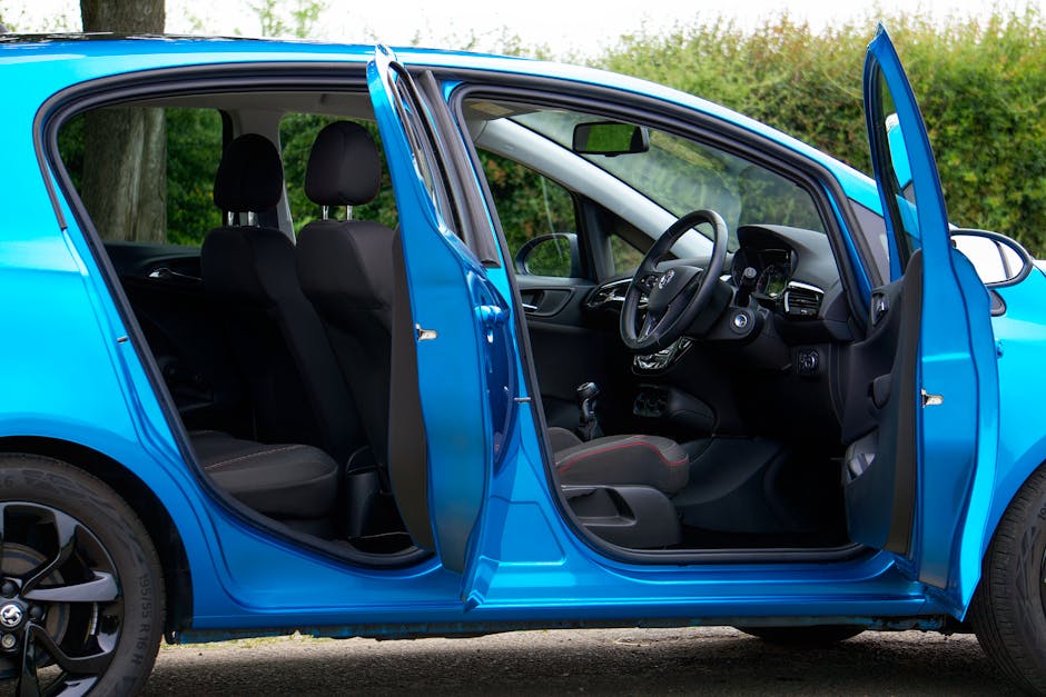 The interior of a blue compact van with its front and rear doors open, revealing the driver's seat, steering wheel, dashboard, and front passenger seat. The rear passenger area contains a row of black fabric-upholstered seats with headrests. On the floor of the rear compartment, various cardboard boxes and plastic-wrapped items are arranged, suggesting packing materials for a home relocation. The van is parked outdoors, with greenery visible in the background, and the scene is illuminated with natural daylight. The loading area shows the process of furniture transport and packing, reflecting logistics typical of professional removals services. Man with Van Vauxhall, a specialist in removals, may utilize such vehicles for efficient furniture transport and household moving jobs, ensuring proper packing and loading during home moves.