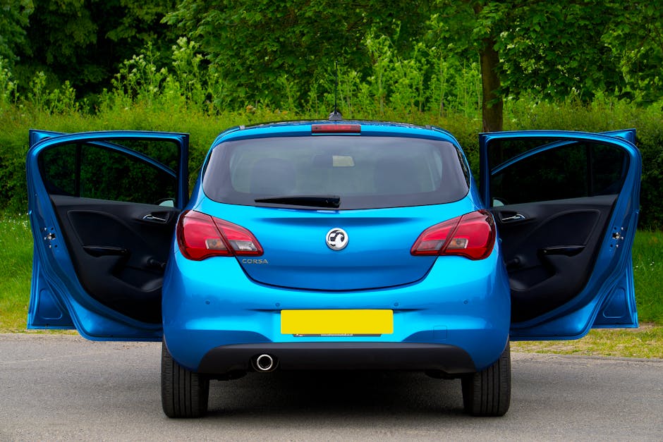 The image shows a bright blue hatchback car with both front doors wide open on a paved road, positioned in front of a lush green background with trees and bushes. Inside, the black interior of the car is visible, with the door panels displaying handles and storage compartments. The rear window has a windshield wiper, and the car's taillights are turned on the sides of the rear. The vehicle is part of a home relocation or furniture transport process, typical in move logistics, and is parked near the edge of a rural or suburban area. The setting suggests an unloading or loading scene, consistent with professional removals services such as those provided by Man with Van Vauxhall, especially as part of relocating household items or furniture. The vehicle's spare wheel is partially visible beneath the rear bumper, and the overall environment appears well-lit with natural daylight, emphasizing the preparation for moving furniture or packed boxes in a house removal context.