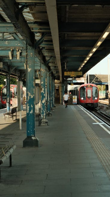 An outdoor train station platform with a metal canopy supported by weathered blue-painted columns. The platform is paved with concrete, featuring tactile paving strips near the edge for safety. A red 