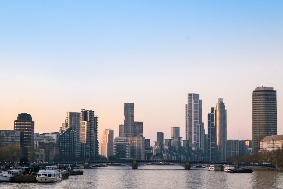 A wide view of a city skyline along a river, featuring numerous modern high-rise office buildings and residential towers with glass facades reflecting natural light. In the foreground, several small boats are moored along the riverbank, and a bridge spans across the water. The scene is captured during daylight without visible human activity. This urban environment illustrates a typical backdrop for house removals and furniture transport services, such as those provided by Man with Van Vauxhall, especially when coordinating home relocation or building access permits in the area around Vauxhall and Lambeth. The image emphasizes the cityscape setting relevant to removals, showing both the logistical environment and the potential context for loading or unloading processes during a house move.