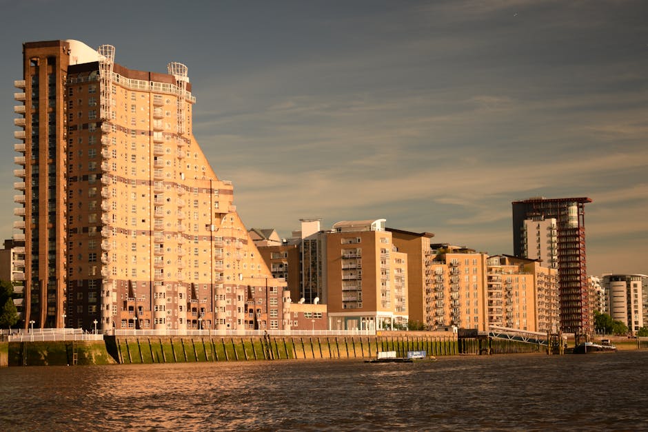 Exterior view of a row of modern high-rise residential buildings with various architectural styles, situated along a riverbank with a concrete retaining wall and grassy slope. The buildings are illuminated by warm sunlight, casting shadows on their facades, and include a large, uniquely shaped tower on the left with rounded balconies and a sloped roof, alongside other apartment blocks with balconies and flat roofs. In the foreground, the river reflects the buildings and the partly cloudy sky above. This scene depicts an urban riverside setting, relevant to home relocation and furniture transport processes, and illustrates the types of properties serviced by Man with Van Vauxhall for removals and house moving services.