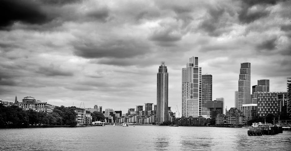 A black-and-white photograph of a city skyline featuring tall modern skyscrapers with varying architectural designs along a river. In the foreground, the river has calm water with a few boats visible, and on the riverbank, there are low-rise residential and commercial buildings. Nearby, there are some trees and open spaces, with the sky above filled with thick, cloudy formations. The scene captures the exterior environment before the house move, illustrating urban moving logistics, with the absence of visible furniture or packing materials, but suggesting the context of home relocation through the city's skyline and river setting. Man with Van Vauxhall, a professional removals company, might be involved in transporting furniture and boxes from here as part of a local move.