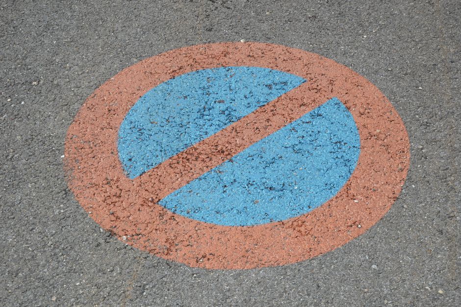 Close-up of a circular road marking on asphalt pavement indicating a no-parking zone, with a blue center and a red border, and a diagonal red line through the middle. The surface features small gravel and asphalt fragments, and the marking appears slightly weathered. This image is relevant to relocation and street management in Vauxhall, as depicted on the webpage about restricted streets and skip permits, and may be part of instructions or signage related to parking restrictions during home removals or furniture transport by Man with Van Vauxhall. The scene is well-lit, showing the marking clearly against the textured pavement.