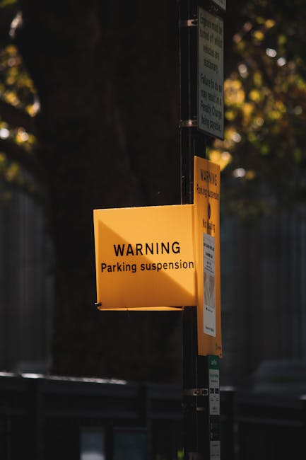 A yellow warning sign attached to a metal pole displays the message 'WARNING Parking suspension' in black text. The sign is situated on a street or urban environment with a dark background, partially obscured by shadows from nearby trees and buildings. Additional signs and information are visible above and below the yellow warning sign, indicating parking restrictions in the area. The scene may be relevant to house removals or relocation services provided by Man with Van Vauxhall, highlighting the importance of understanding local parking regulations during furniture transport and home moving processes.
