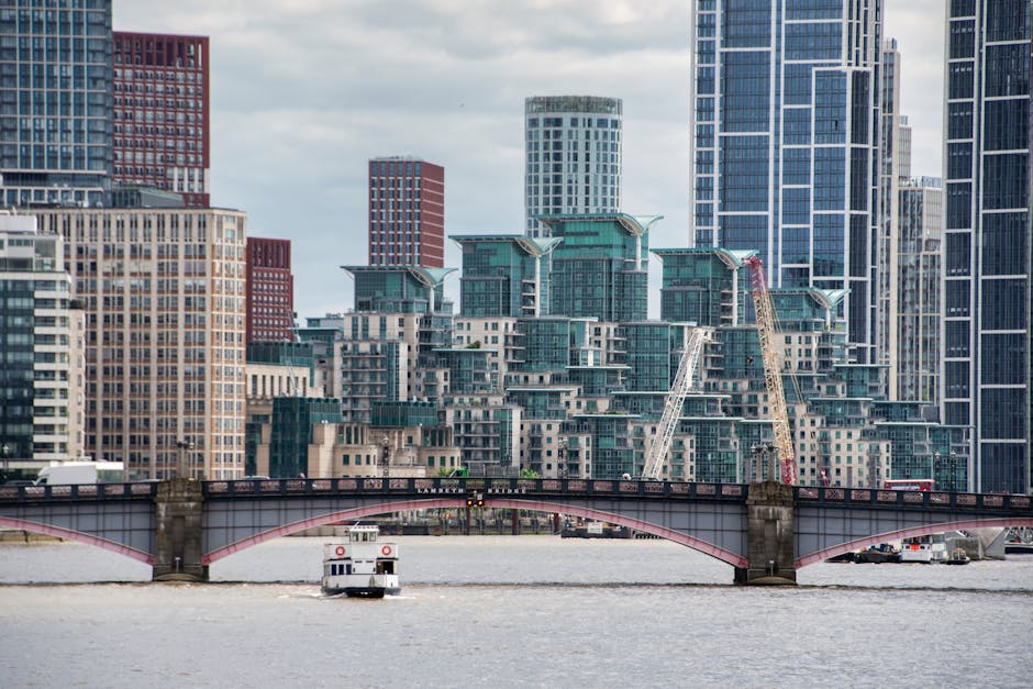 A daytime cityscape featuring a river in the foreground with a small passenger ferry navigating the water. Behind the river, there is a multi-story bridge with a red and gray structure spanning across