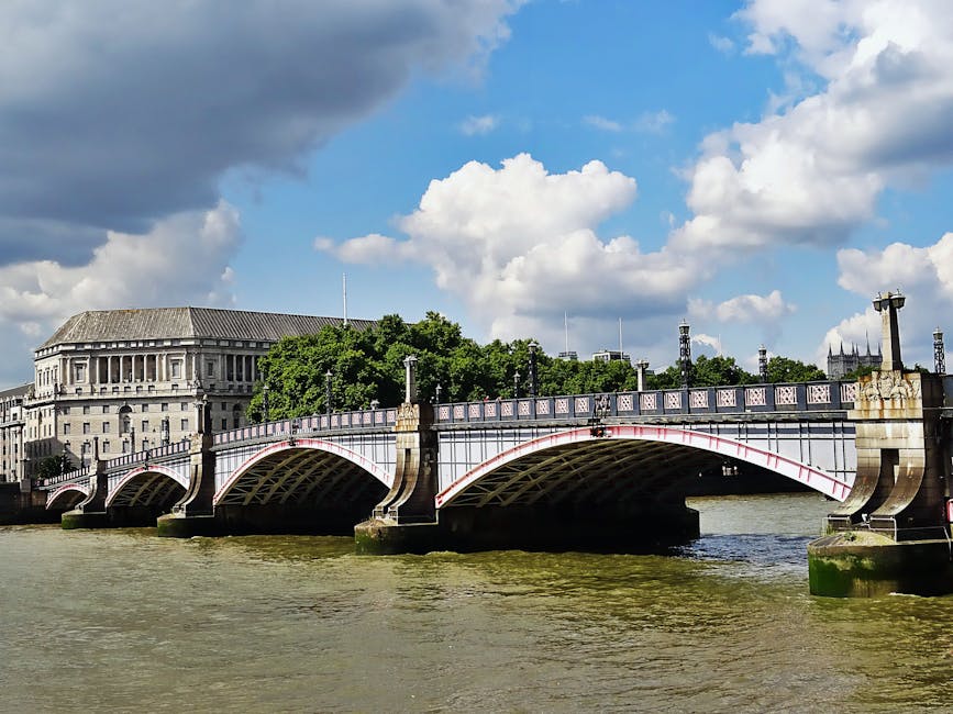 A daytime cityscape featuring a river in the foreground with a small passenger ferry navigating the water. Behind the river, there is a multi-story bridge with a red and gray structure spanning across